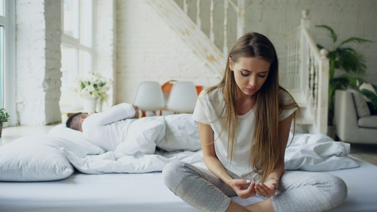 Woman sitting on bedroom floor looking thoughtful and reflective while partner sits on the bed, symbolising emotional reflection and personal boundaries.