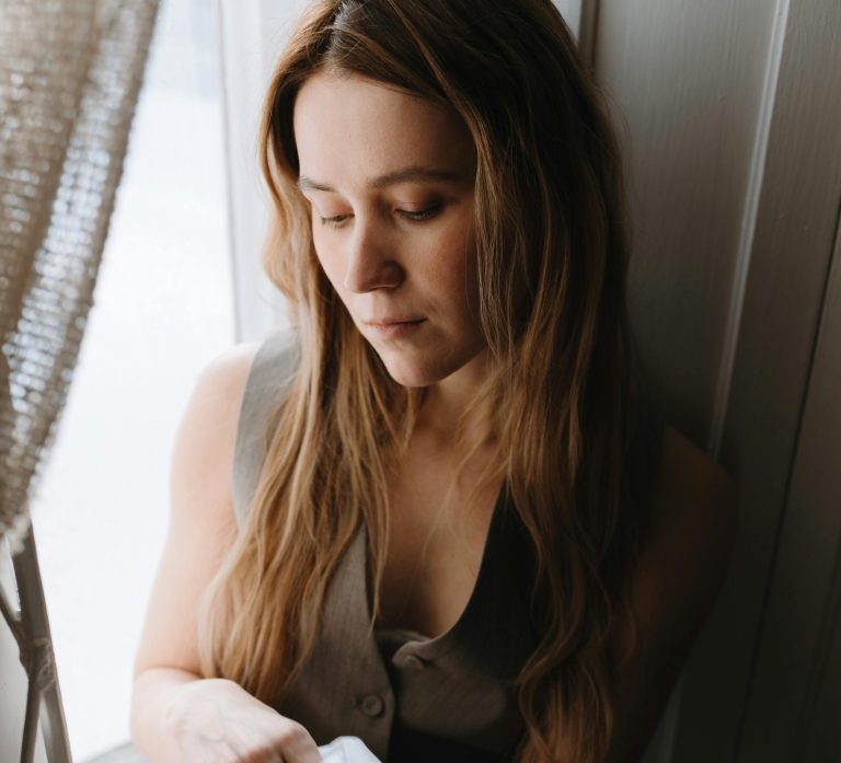 Woman looking out the window, appearing reflective and pensive, symbolising life transitions and feeling stuck.