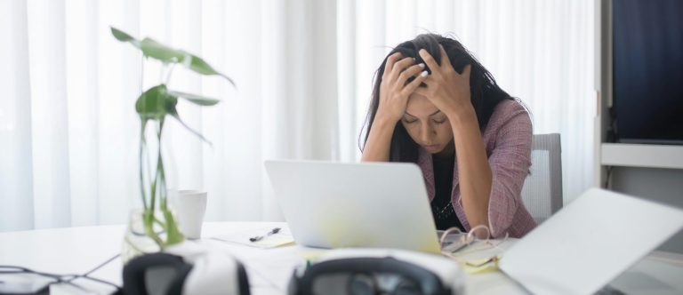 Woman sitting at desk with head in hands, feeling exhausted and stressed from work, showing subtle signs of burnout in women.